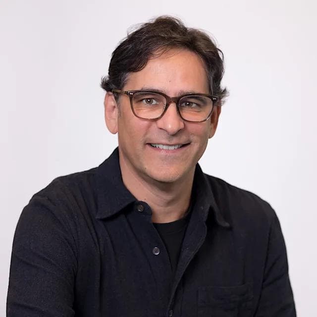 Smiling man with dark hair and glasses wearing a black shirt against white background.
