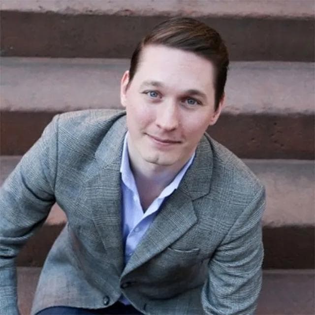 Smiling man with blue eyes wearing a grey plaid blazer sitting on stone steps.