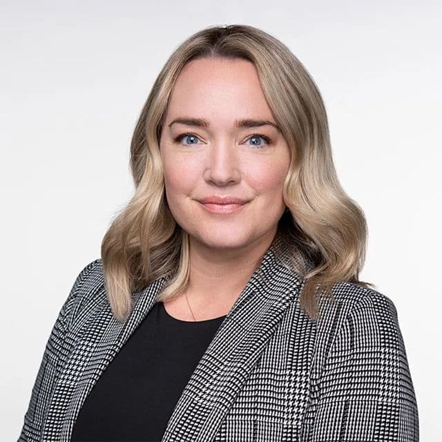 Blonde woman with blue eyes wearing a black and white patterned blazer against white background.